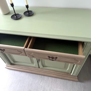 May include: A light green painted wooden cabinet with two drawers open, revealing a green interior. The cabinet has a light beige base and drawer fronts with ornate handles. Two black candle holders with white candles and a white pitcher are on top.
