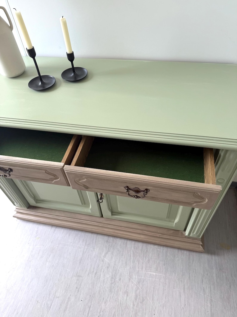 May include: A light green painted wooden cabinet with two drawers open, revealing a green interior. The cabinet has a light beige base and drawer fronts with ornate handles. Two black candle holders with white candles and a white pitcher are on top.
