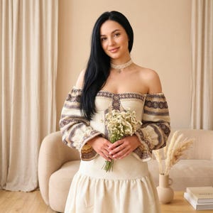 May include: A woman wearing a cream-colored off-the-shoulder dress with brown and beige embroidered details. The dress has long sleeves and a fitted bodice. She is holding a bouquet of white flowers and wearing a lace choker.