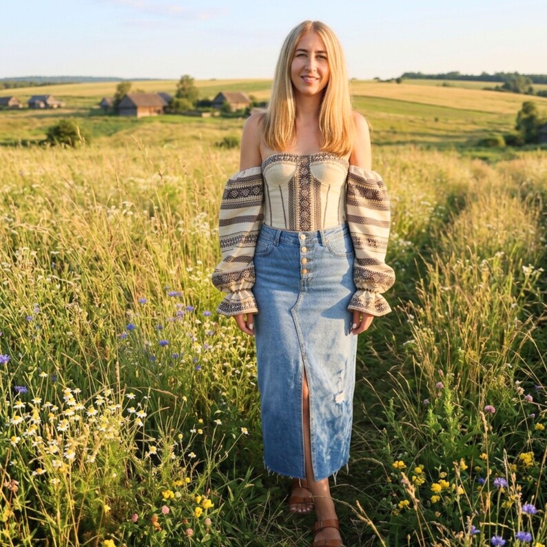 May include: A woman wearing a beige off-the-shoulder top with black and white patterned sleeves and a denim skirt. The skirt has a front slit and button details. The outfit is worn outdoors in a field of tall grass.