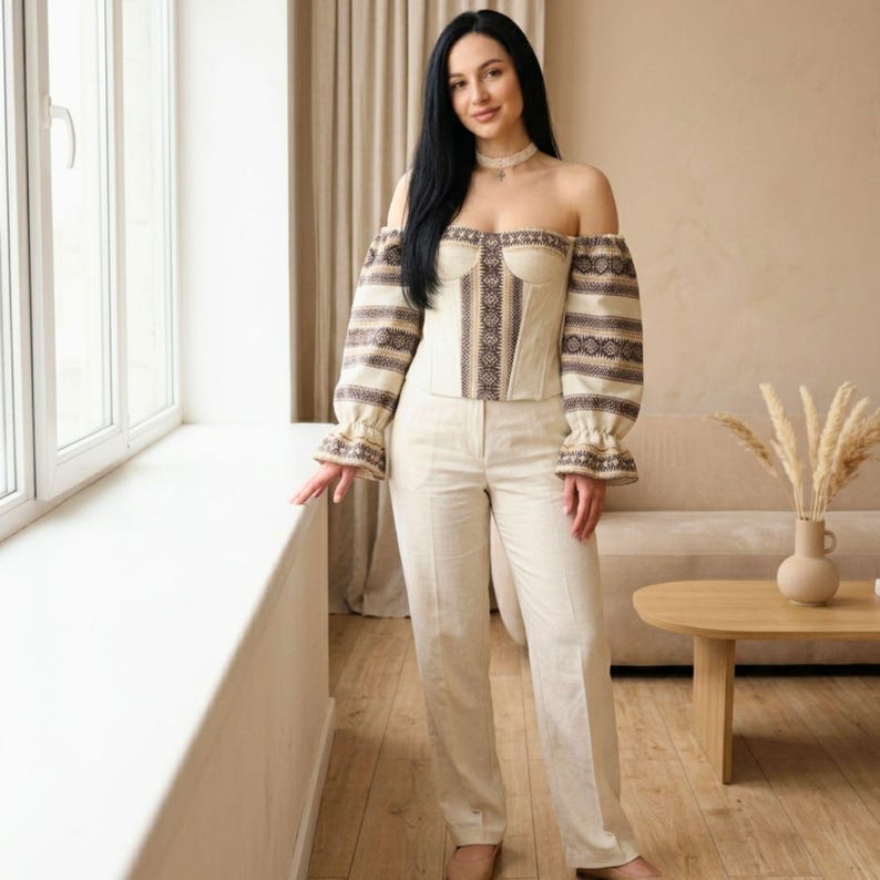 May include: Off-the-shoulder cream-colored top and matching pants. The top features intricate embroidery and long sleeves with ruffled cuffs. The pants are high-waisted with a straight leg. The outfit is made of a light-colored fabric.