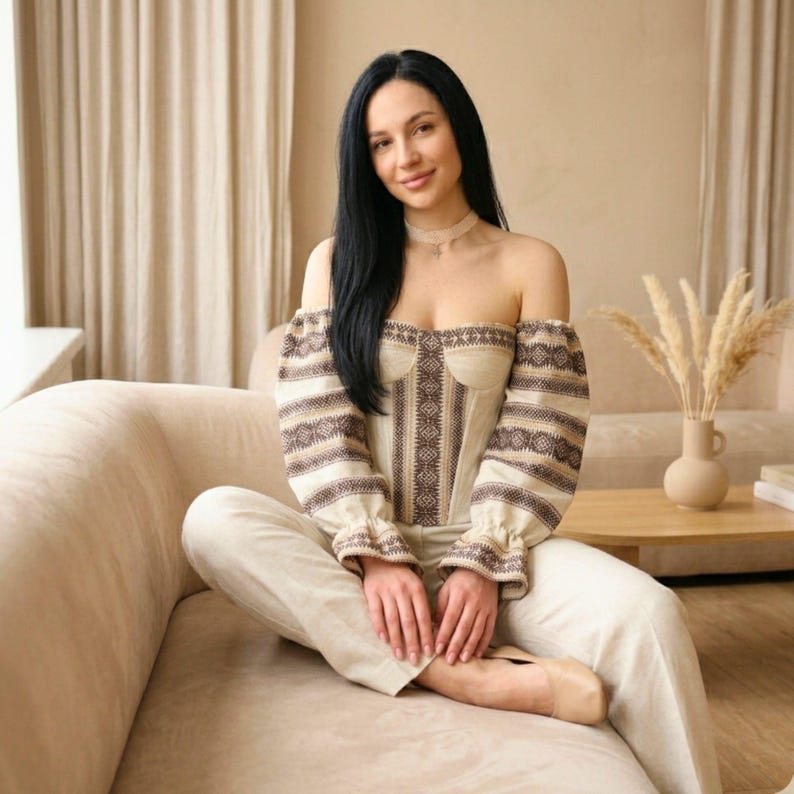 May include: Off-the-shoulder beige corset top with brown embroidered details and long sleeves. The top is paired with matching beige pants. The woman is sitting on a beige sofa.