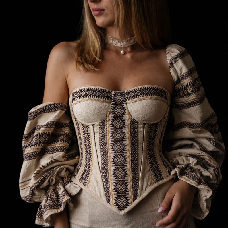 May include: A beige corset with intricate brown and blue geometric patterns. The corset features a strapless design with one long sleeve and a fitted bodice. The garment is part of a traditional clothing ensemble.