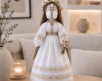 Puede incluir: Muñeca hecha a mano con un vestido blanco con bordados beige y una corona floral. La muñeca tiene pelo largo castaño y sostiene un pequeño ramo. El vestido tiene mangas largas y una cintura detallada. Es un artículo decorativo.