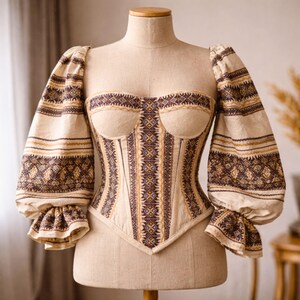 May include: A beige corset top with long, puffed sleeves. The bodice and sleeves feature intricate geometric patterns in brown and gold. The corset is displayed on a mannequin.