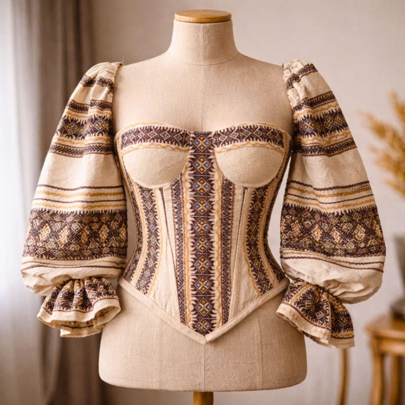 May include: A beige corset top with long, puffed sleeves. The bodice and sleeves feature intricate geometric patterns in brown and gold. The corset is displayed on a mannequin.