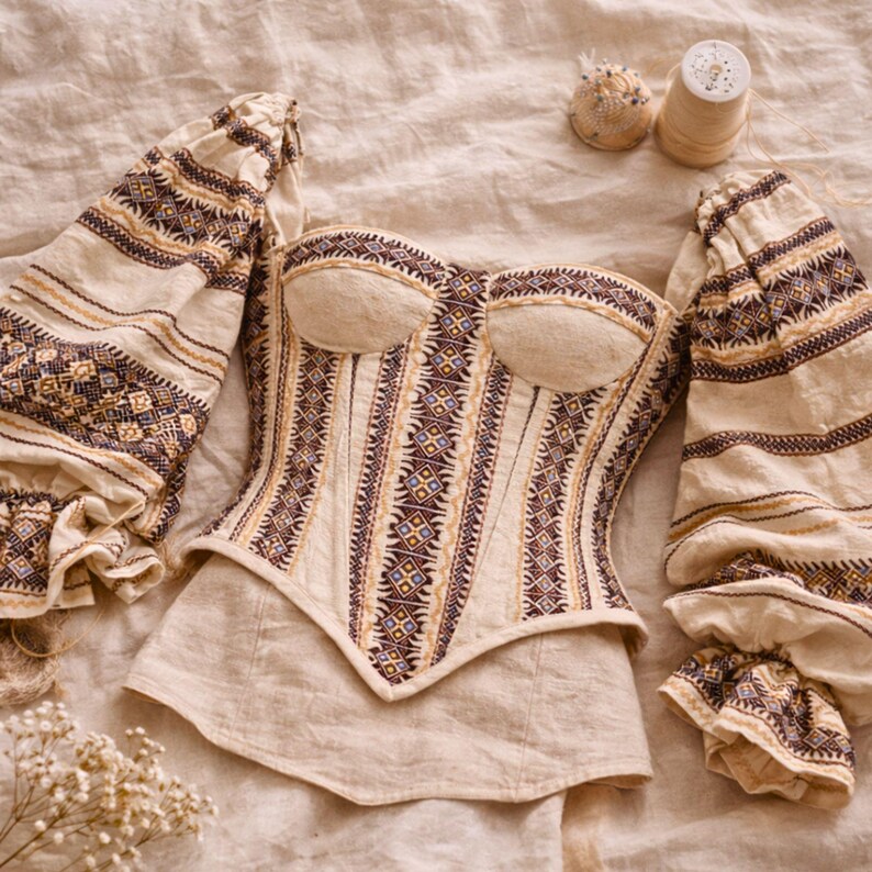 May include: Cream-colored corset and sleeves with intricate embroidered patterns in brown, gold, and blue. The corset features a structured bodice and a sweetheart neckline. The sleeves are long and puffy, with matching embroidery. The ensemble is laid out on a neutral-toned fabric.