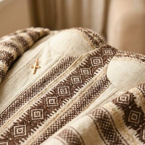 May include: Close-up of a beige garment with intricate brown geometric embroidery and a gold-colored cross pin. The fabric has a textured appearance, and the design features repeating diamond patterns.