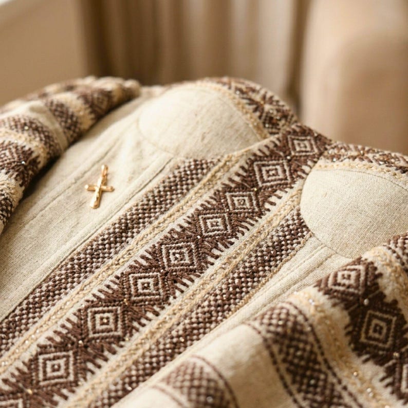 May include: Close-up of a beige garment with intricate brown geometric embroidery and a gold-colored cross pin. The fabric has a textured appearance, and the design features repeating diamond patterns.