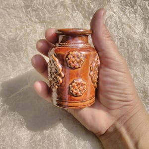 Op de afbeelding: Een kleine, bruine keramische pot met ingewikkelde reliëfmotieven. De pot heeft een rond lichaam en een licht uitlopende bovenkant.