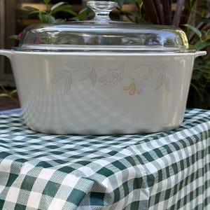 May include: A white casserole dish with a clear glass lid. The dish has a floral design in soft pink and yellow. The dish is sitting on a green and white checkered tablecloth.