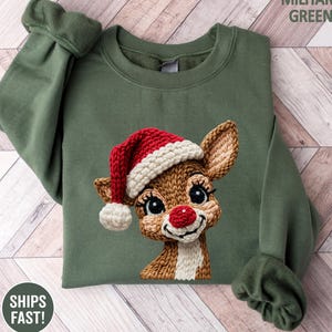 May include: Military green sweatshirt featuring a knitted reindeer wearing a Santa hat. The reindeer has a red nose and is smiling. The sweatshirt has long sleeves and a crew neck. The text "MILITARY GREEN" is in the upper right corner.