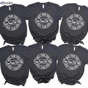 Könnte beinhalten: Sechs dunkelgraue T-Shirts mit einem weißen Kreisdesign, auf dem "BEST. TEAM. EVER" und "Team Name/Job Title" in der Mitte stehen.