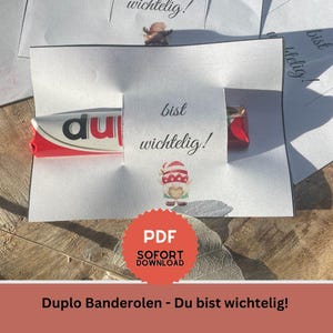 May include: A white paper wrapper with the text "bist wichtig!" surrounds a Duplo candy bar. A small gnome sticker and a red "PDF SOFORT DOWNLOAD" badge are also visible. The bottom of the image has the text "Duplo Banderolen - Du bist wichtig!"