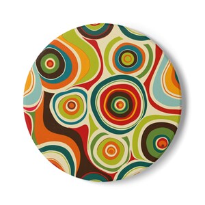 Op de afbeelding: Een ronde onderzetter met een retro design. De onderzetter heeft een patroon van kleurrijke concentrische cirkels in tinten oranje, groen, blauw, rood en bruin tegen een crèmekleurige achtergrond. Het ontwerp doet denken aan de jaren 1960 en 1970.