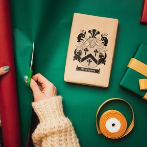 May include: A person wrapping a gift with green wrapping paper. The gift is a brown box with a black and white coat of arms and the text "Davenport". The person is using a pair of scissors to cut the wrapping paper. There is a roll of red wrapping paper and a gold ribbon on the table.