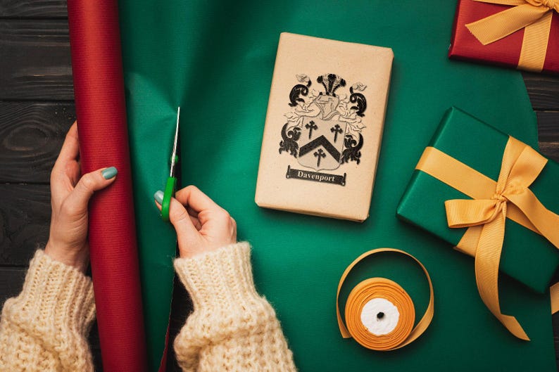 May include: A person wrapping a gift with green wrapping paper. The gift is a brown box with a black and white coat of arms and the text "Davenport". The person is using a pair of scissors to cut the wrapping paper. There is a roll of red wrapping paper and a gold ribbon on the table.