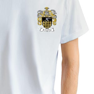 May include: A white t-shirt with a crest featuring a black and yellow checkered shield with a white bird on top. The crest is surrounded by a gold and silver floral design. The text "Antolini" is written in a black banner below the crest.