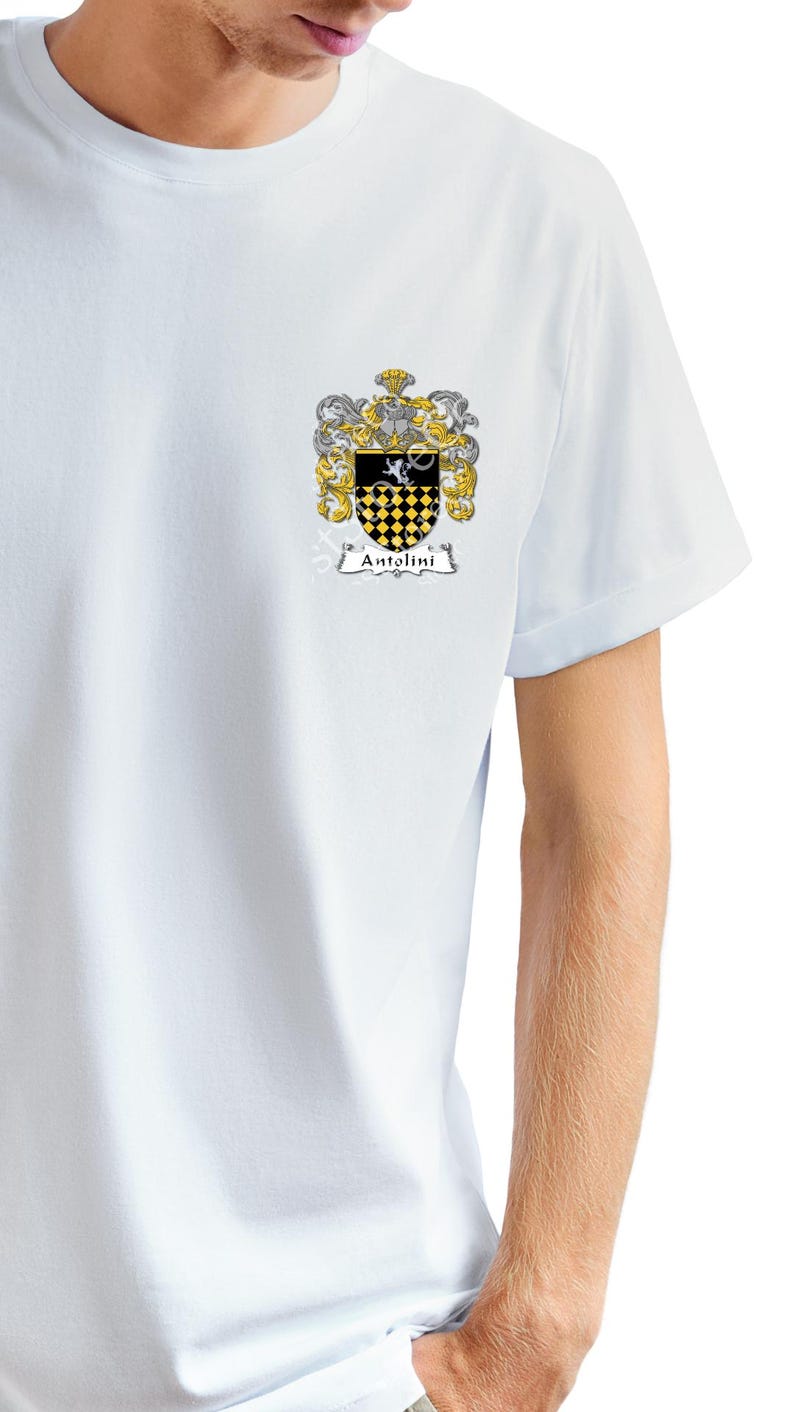 May include: A white t-shirt with a crest featuring a black and yellow checkered shield with a white bird on top. The crest is surrounded by a gold and silver floral design. The text "Antolini" is written in a black banner below the crest.
