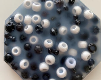 May include: An octagonal resin coaster with a mix of white and black beads embedded within a translucent, light blue-grey background. The beads vary in size and shape, creating a textured surface.