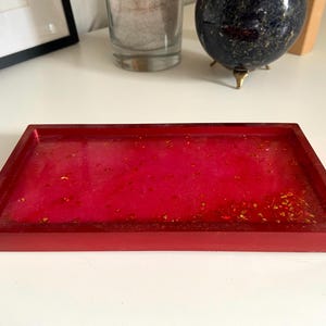 May include: A rectangular red resin tray with a glossy finish. The tray's interior is a vibrant pink with gold flecks. The tray is a decorative item, suitable for holding small items.