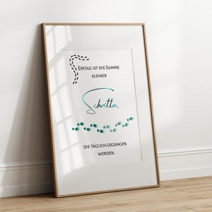 Könnte beinhalten: Gerahmter Druck mit einem motivierenden Zitat auf Deutsch. Der Text lautet: "Erfolg ist die Summe kleiner Schritte, die täglich gegangen werden." Das Design umfasst schwarze gepunktete Linien und türkisgrüne Fußabdrücke. Der Rahmen ist hellbraun.