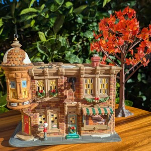 May include: A detailed ceramic miniature of a two-story building with a pharmacy sign and a welcome mat. The building is made of brick and has a green and white striped awning. A small tree with orange leaves is in the background.