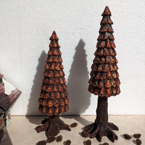May include: Two brown decorative pine trees with detailed bark and cone textures. The trees have a rustic appearance and are surrounded by small pine cones, suggesting a woodland theme. The trees are likely for seasonal decor.