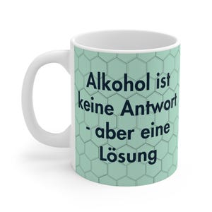 Può includere: Tazza in ceramica bianca con un motivo a nido d'ape verde chiaro e il testo tedesco "Alkohol ist keine Antwort - aber eine Lösung" in nero. La tazza ha un manico curvo e una forma cilindrica.