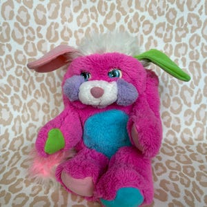 Peut inclure: Une peluche en forme de lapin rose vif, avec des accents bleus, verts et violets. Il a de grands yeux bleus expressifs, de longues oreilles et une queue touffue. Le jouet est conçu avec un tissu doux et texturé, et est un jouet pour enfants.