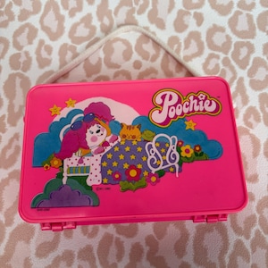 May include: A bright pink vintage lunchbox with a cartoon graphic of a girl, cat, and stars. The lunchbox has a white handle and the word "Poochie" in a cursive font. The background is a pink and white leopard print.