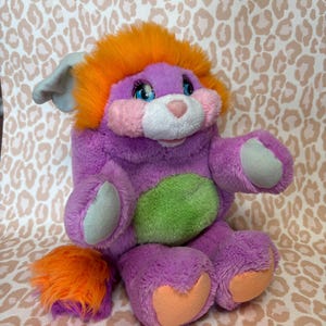 May include: A plush toy featuring a purple body with a bright orange mane and tail, a light green belly patch, and pink cheeks. The creature has large blue eyes, a white muzzle, and light blue accents on its ears, paws, and feet.