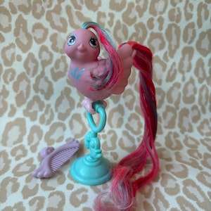 Op de afbeelding: Een roze vogelvormig speelgoed met een lange, kleurrijke staart en blauw en roze haar. Het speelgoed is gemonteerd op een lichtblauwe standaard en bevat een kleine, lavendelkleurige kam. De vogel heeft blauwe markeringen en grote ogen.