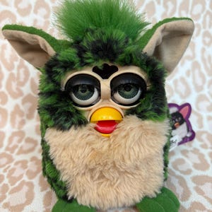 Może przedstawiać: Zabawka Furby w stylu vintage z zielonym i czarnym futrem w kamuflażu, beżową klatką piersiową i zielonymi, puszystymi włosami. Ma duże, wyraziste oczy, żółty dziób i czerwony język. Zabawka jest przedmiotem kolekcjonerskim.