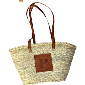 Peut inclure: Sac fourre-tout en paille tressée avec anses en cuir marron et un patch carré en cuir marron. Le patch porte la lettre "P". Le sac a un large dessus ouvert et est tenu par des anses en cuir marron.