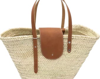 May include: A large, woven straw tote bag with brown leather handles and a brown leather flap. The bag has a wide, open top and a rounded bottom.