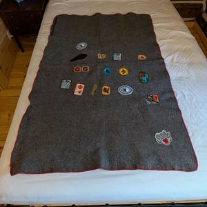 Op de afbeelding: Een donkergrijze wollen deken met een rode rand, versierd met diverse geborduurde patches. De patches tonen kleurrijke ontwerpen en tekst, waaronder een "Scout Camp" patch. De deken ligt op een wit bed.