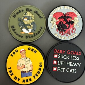 May include: Four circular morale patches. Top left: olive green with a penguin in tactical gear and text "Wake Up And Choose Violence". Top right: light blue with a red heart, mummy figures, and "TRAUMA BONDING SINCE 1775". Bottom left: yellow with a man holding a drink and "YOU CAN TAX MY ASS FEDBOI". Bottom right: black with "DAILY GOALS" and a checklist for "SUCK LESS", "LIFT HEAVY", and "PET CATS".