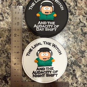 Op de afbeelding: Twee ronde patches met een stripfiguur met een headset en een groene jas. Eén patch is zwart met de tekst "THE LION, THE WITCH AND THE AUDACITY OF DAY SHIFT." De andere is wit met "NIGHT SHIFT."