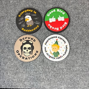 May include: Four round patches with humorous designs. One patch features an eagle and the text "Everyday is Sadderday." Another says "Good Bird Broke Bird" with thumbs up. A third patch has a skull and the words "Defund Operations." The last says "Maintenance Control, Your Ultimate Party Planners."