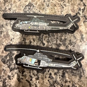 May include: Two black and grey helicopter patches. The patches feature a detailed illustration of a military helicopter with the word "MARINES" printed on the side. The patches are on a speckled brown surface.
