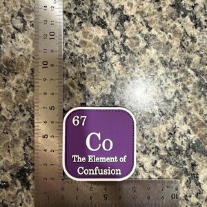 以下が含まれることがあります： 「67 Co The Element of Confusion」の白文字が入った正方形の紫色のパッチ。 パッチの幅は約5cmで、定規の隣に配置されています。