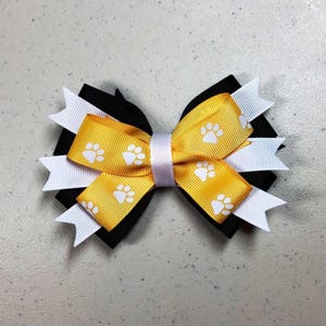 May include: A handmade hair bow featuring a layered design with black, white, and yellow ribbons. The yellow ribbon is printed with white paw prints. A white ribbon wraps around the center of the bow.