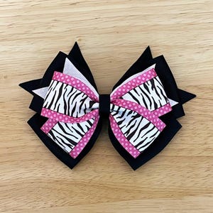 May include: A layered hair bow featuring a zebra print design with pink polka dot accents. The bow has a black base with white and pink ribbon layers. The bow is set against a wooden background.