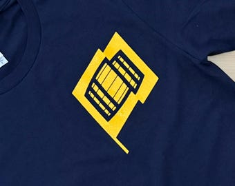 May include: A dark blue t-shirt featuring a yellow graphic print on the chest. The design consists of a yellow diamond shape containing a black outline of a building with multiple windows and a central rectangular structure.