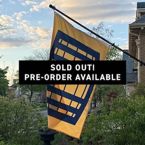 May include: A rectangular yellow flag featuring a dark blue geometric grid pattern, mounted on a black pole attached to a brick wall. The flag displays the text SOLD OUT! PRE-ORDER AVAILABLE in white letters on a black rectangular background.