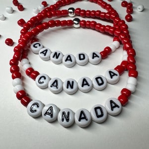 Könnte beinhalten: Drei kreisförmige Armbänder aus kleinen roten und weißen Perlen. Jedes Armband enthält weiße runde Perlen mit dem schwarzen Aufdruck CANADA. Eine einzelne metallisch silberne Perle befindet sich oben an jedem Armband zwischen den roten Perlen.