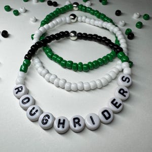 May include: Stack of beaded bracelets in white, green, and black. One bracelet spells out "ROUGHRIDERS" in black letters on white beads. The other bracelets are solid colors with silver beads.