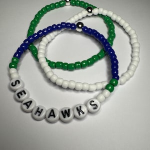 Pode incluir: Três pulseiras de contas verdes, brancas e azuis. Uma pulseira soletra "SEAHAWKS" em contas de letras brancas. Cada pulseira tem um detalhe de contas prateadas.