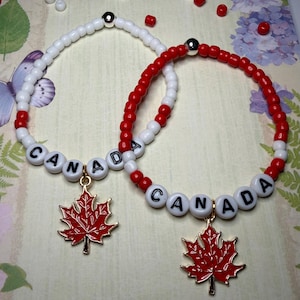 May include: Two elastic bracelets featuring white and red seed beads with a silver-toned spacer bead. Each bracelet includes white letter beads spelling CANADA and a red enamel maple leaf charm with a gold-toned metal border and attachment loop.